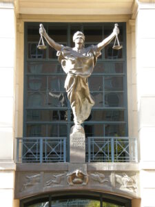 A statue of a blindfolded woman holding scales atop a plaque that reads Justice Delayed Justice Denied