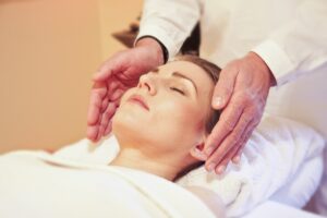 A woman lying on a table with her eyes closed while someone wearing white holds their hands around an inch from her face on both sides of her head